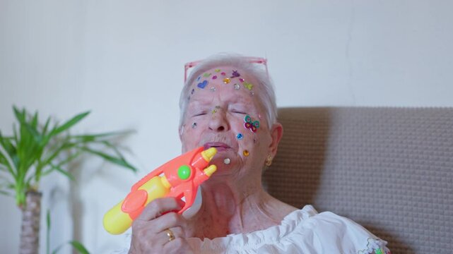 Playful elderly woman with her face decorated with colorful stickers pointing and shooting a water gun at the camera. Embodying a youthful spirit and the joy of embracing one's inner child at any age