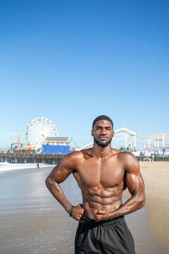 Man flexing muscles on a sunny beach with a pier in the background.