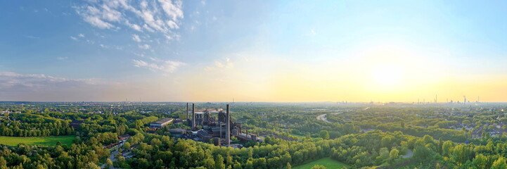 Obraz premium Eine beeindruckende Luftaufnahme des Landschaftsparks Duisburg-Nord zeigt die stillgelegten Hochöfen und Schornsteine des alten Hüttenwerks, umgeben von dichten, grünen Baumkronen.