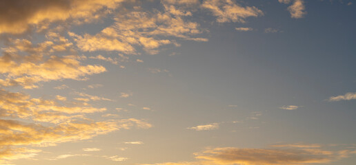 Obraz premium Sunset sky background with golden orange clouds scattered across a soft blue sky. Warm dramatic cloudscape panorama, nature background..