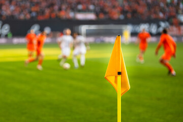 A close-up of a bright yellow corner flag on a grass pitch with a football match and stadium crowd blurred in the background. © Dziurek