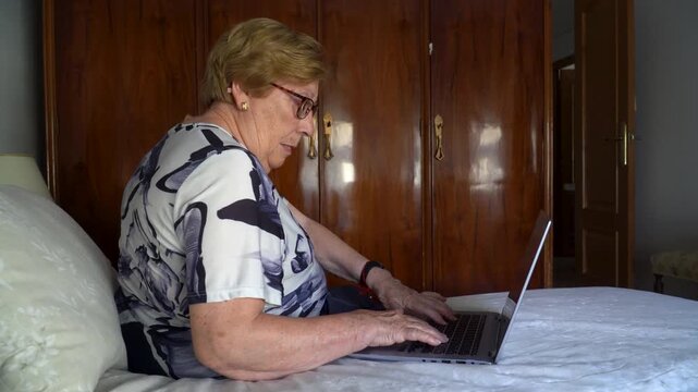  Senior woman using a laptop sitting on the bed at home.