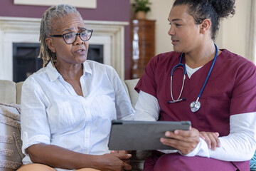 Obraz premium Senior African American woman sitting on couch talking with nurse in maroon scrubs holding tablet