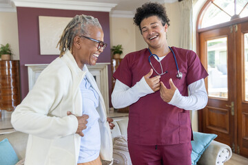 Obraz premium Male caregiver in burgundy scrubs, stethoscope gesturing with Senior African American woman at home