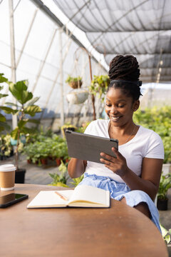 African American woman sitting at wooden table in greenhouse holding tablet smiling notebook nearby