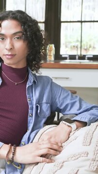 Vertical video: Shifting nonbinary adult in maroon glancing left, tucking curl on couch by window