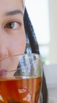 Vertical video: Lifting glass of tea, woman sipping at kitchen window, headband, plant, copy space