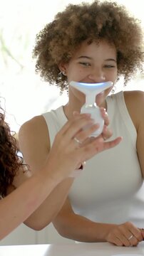 Vertical video: Picking LED device, left woman guiding tip along cheek, testing product at counter