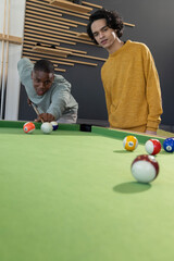 Obraz premium African American and Asian men playing pool in lounge on green-felt table, aiming among pool balls