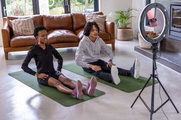 Obraz premium Diverse duo in activewear stretching on green mats in living room, recording with ring light phone