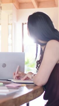 Vertical video: Writing woman in maroon top glancing at window glare and noting in notebook at home