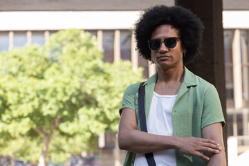 Fototapeta premium African American man standing right in sunny plaza, wearing green-shirt, shades, bag, copy space