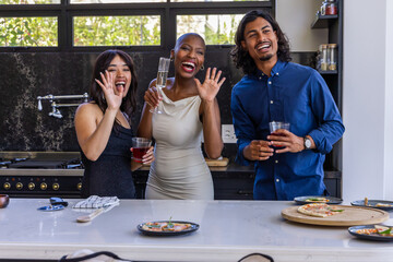 Diverse friends waving and holding drinks behind kitchen island, showing pizza slices and flute © wavebreak3