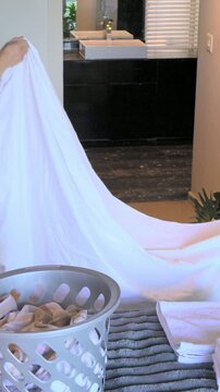 Vertical video: Grasping corners, 2 hands pulling white sheet across bed, making bed with basket