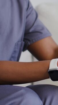 Vertical video: Checking watch, carer in scrubs holding senior hand for support on sofa, copy space