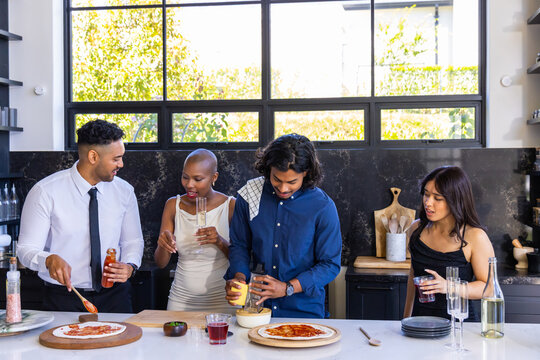 Diverse friends gathering at kitchen island preparing pizzas on wooden boards with jar, grater
