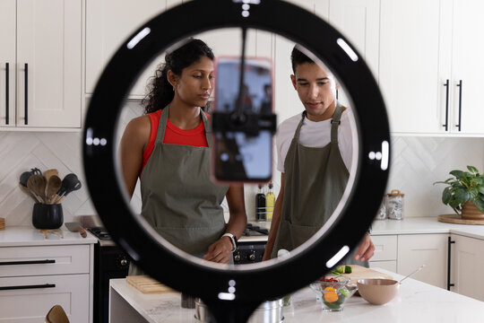 African American couple in gray aprons cooking and recording using ring light and phone in kitchen