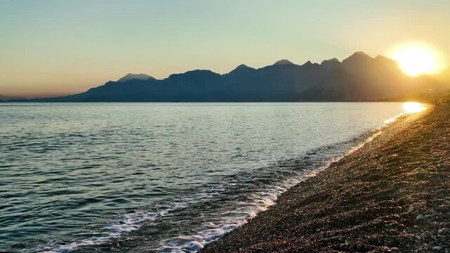 Sun sets behind taurus mountains casting golden light over calm mediterranean sea and pebble beach.