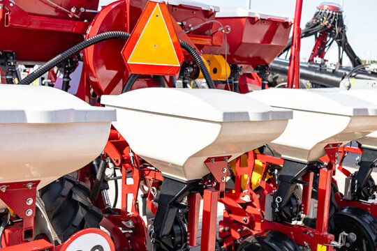 Detailed close-up view of red row-crop planting equipment showing white seed hoppers, metal frames, wheels and drive components under bright outdoor light on