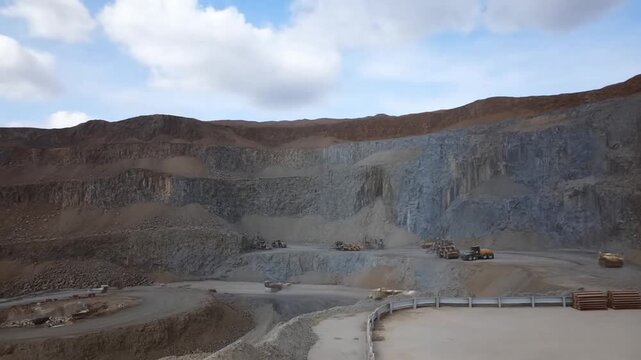 Aerial tracking shot of dump trucks hauling material and loaders working in open pit quarry with terraced rock walls and cloudy sky