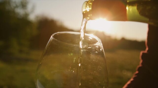 A hand pours a radiant white wine into a glass as sunlight dances through the liquid, capturing the essence of a luxurious evening at a vineyard