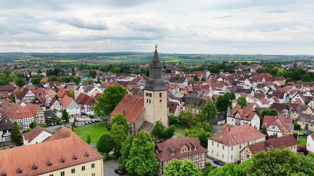 Blick &uuml;ber die D&auml;cher der historischen Altstadt von Hofgeismar mit der markanten Altst&auml;dter Kirche im Zentrum, umgeben von gr&uuml;nen B&auml;umen und sanften H&uuml;geln unter bew&ouml;lktem Himmel.