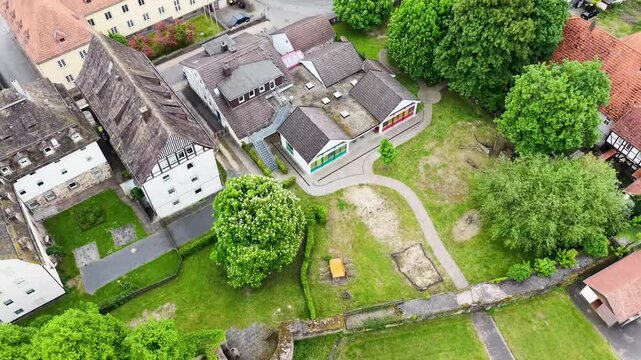 Blick &uuml;ber die D&auml;cher der historischen Altstadt von Hofgeismar mit der markanten Altst&auml;dter Kirche im Zentrum, umgeben von gr&uuml;nen B&auml;umen und sanften H&uuml;geln unter bew&ouml;lktem Himmel.