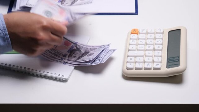 Person is counting us hundred dollar bills by hand next to a calculator on a white desk, managing personal finance, budgeting, and calculating expenses