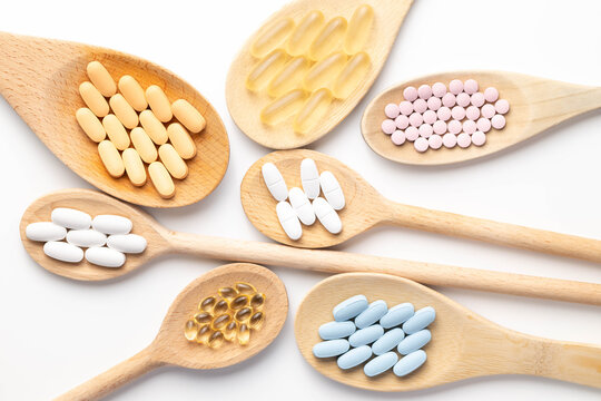 Wooden spoons holding various health supplements, soft gels, capsules, tablets, on white background.