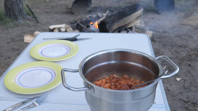 Stainless steel pot filled with warm baked beans for an outdoor camping meal, placed on a folding table with empty plates and cutlery, a burning campfire glowing in the background
