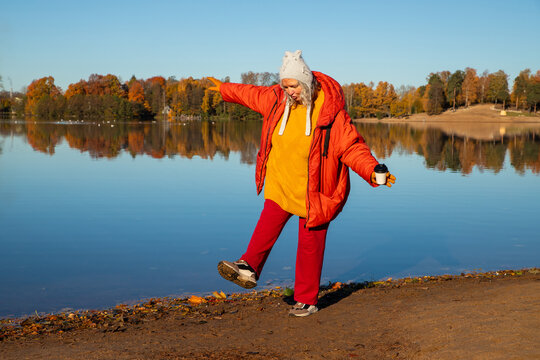 47 year old body positive woman in orange jacket walking near lake in autumn. lady relaxing outdoor in nature with coffee. Lifestyle concept of enjoying fall season during weekend.
