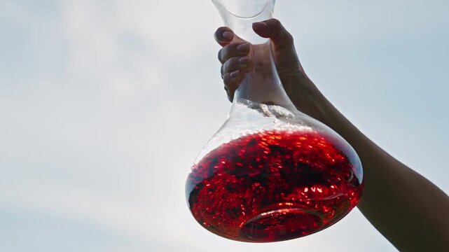Woman Swirling Red Wine in Crystal Decanter Against Sunlit Sky Background