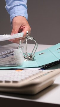 Vertical video employee hand placing plastic sleeves with documents into an open ring binder on a desk, next to a calculator, managing business files and financial records