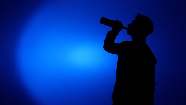 Male silhouette drinking from a bottle against a deep blue background, conveying alcoholism, addiction, isolation and dependence in a stark, moody scene