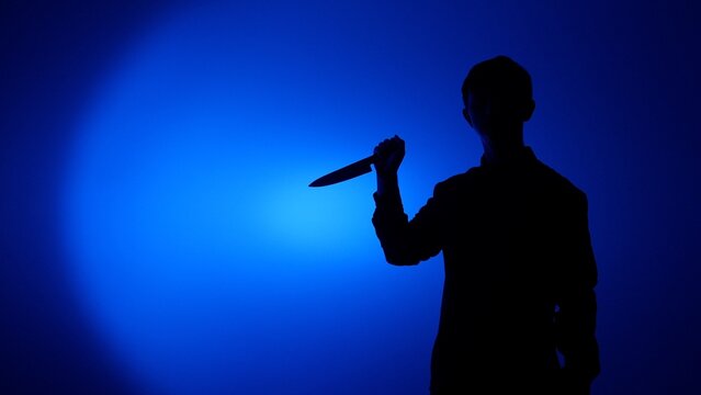 Silhouette of a man holding a sharp knife in dramatic blue backlight and spotlight, evoking danger, threat, mystery and violence with concealed identity and ominous mood