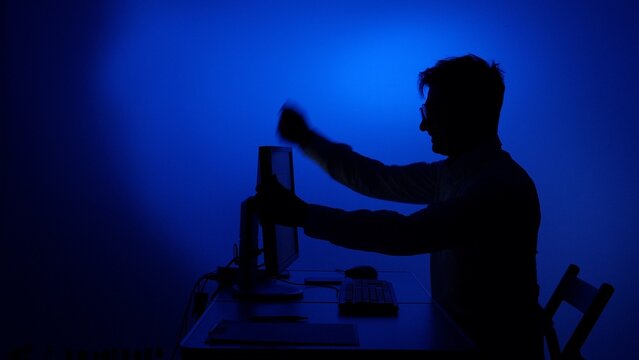 Man feeling frustrated and angry, hitting a computer monitor on a desk, dealing with emotional stress and burnout in a dark, blue lit environment, symbolizing technological issues