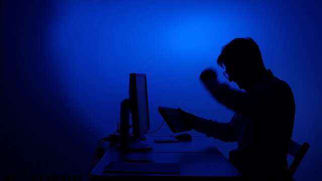 Silhouette of a frustrated person in glasses showing strong anger while violently grasping the keyboard and working on a computer against a blue background, representing digital stress