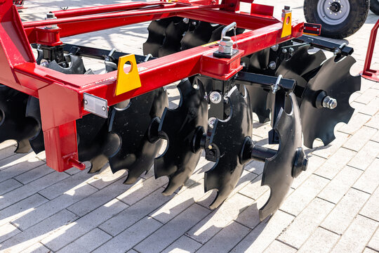 Close-up view of a red disc harrow implement mounted on a farm trailer, showing rows of black concave steel discs, a sturdy red frame with yellow safety