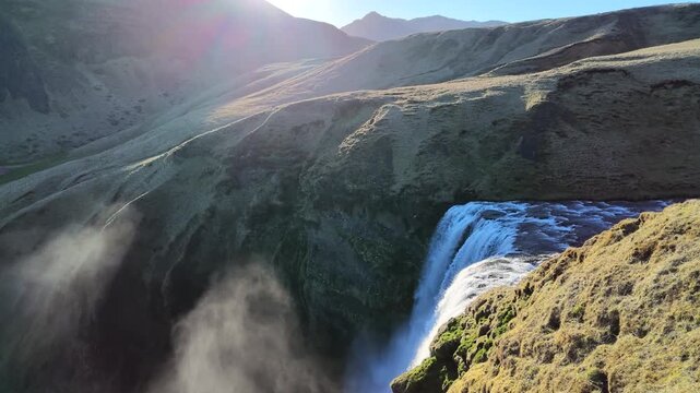 puissante cascade islandaise plongeant dans un canyon verdoyant. Le soleil &eacute;claire la sc&egrave;ne tandis que les embruns s'&eacute;l&egrave;vent au-dessus des falaises.