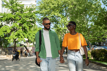 Naklejka premium Cheerful african american couple walks with hold hands in sidewalk downtown, guy makes girlfriend laugh by fun storytelling. Stylish black young woman enjoy stroll with happy boyfriend.