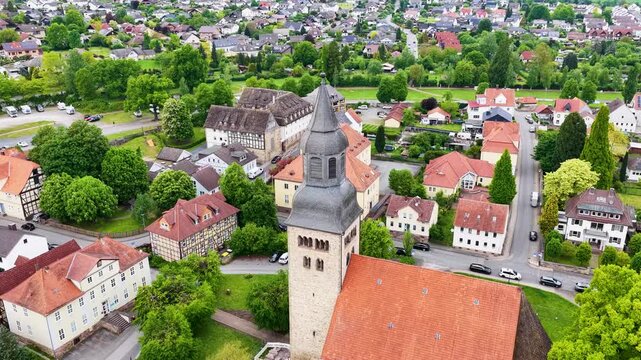 Blick &uuml;ber die D&auml;cher der historischen Altstadt von Hofgeismar mit der markanten Altst&auml;dter Kirche im Zentrum, umgeben von gr&uuml;nen B&auml;umen und sanften H&uuml;geln unter bew&ouml;lktem Himmel.