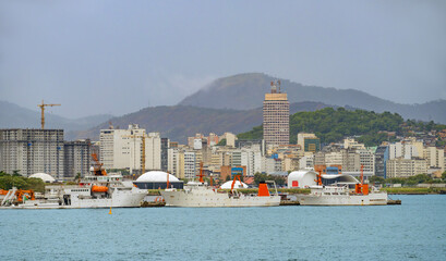 Several white industrial ships are docked in a harbor. Behind them stands a city skyline featuring a tall tower and buildings under construction. Hazy mountains rise in the background © John_Doo78