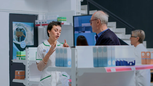 Aged client in need of prescription illness prevention drug information in pharmacy, asking knowledgeable pharmacist, with pharmacy employee helping older customer with medical expertise.