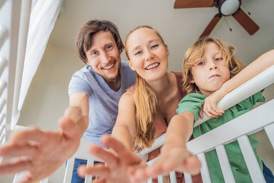 Parents and older brother looking into baby crib from infant point of view. Family smiling down at newborn with love and care. Sibling bonding, parenthood, childhood memory and connection concept