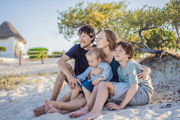 Fototapeta premium Happy family with mother, father and two sons standing on the beach among tropical coastal bushes. Summer vacation, parenting, travel and family bonding concept by the sea