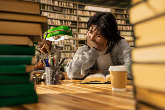 Tired young woman studying open book under pressure at library desk, showing academic fatigue, stress and frustration during coursework preparation, finals burnout, for study concepts.