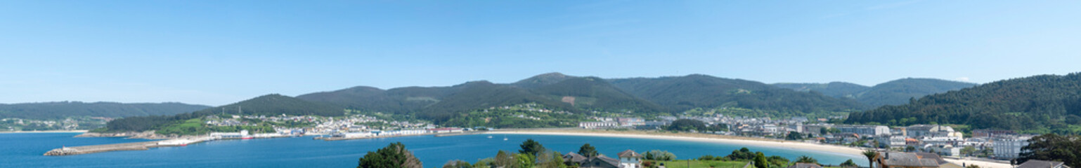 Fototapeta premium A sweeping panoramic view of Viveiro town curved around the sandy bay and blue sea of the Covas beach with rolling green mountains under a clear sky in Galicia, Spain.