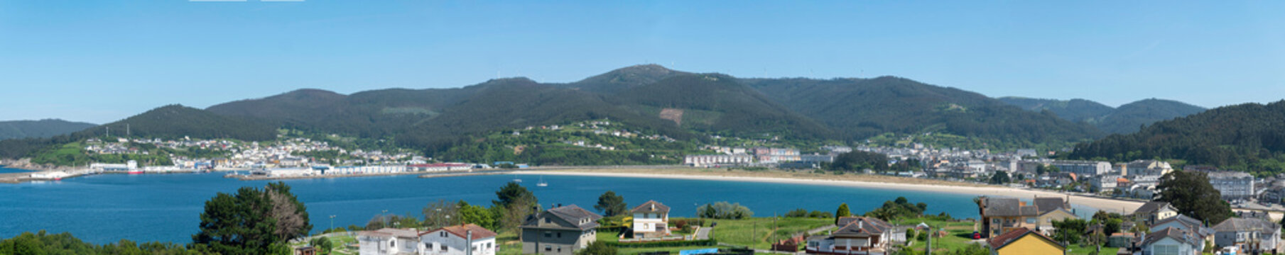 A sweeping panoramic view of Viveiro town curved around the sandy bay and blue sea of the Covas beach with rolling green mountains under a clear sky in Galicia, Spain.