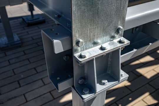 Close-up view of a galvanized steel support bracket bolted to an outdoor metal framework over a paved surface. Visible hex bolts, welded seams, and textured