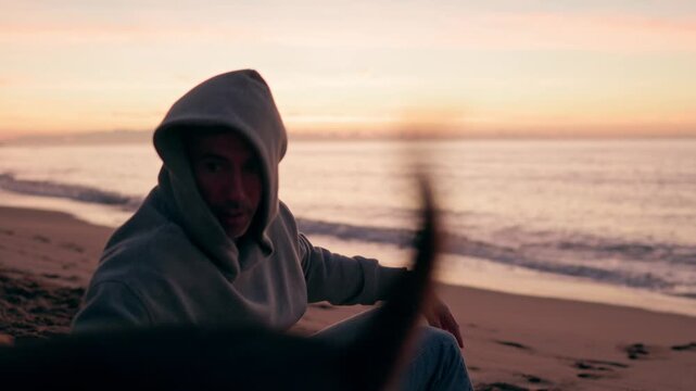 Melancholic man in a hoodie sitting on a sandy beach at sunset, petting his dog while looking towards the camera with a thoughtful expression as the waves gently roll in